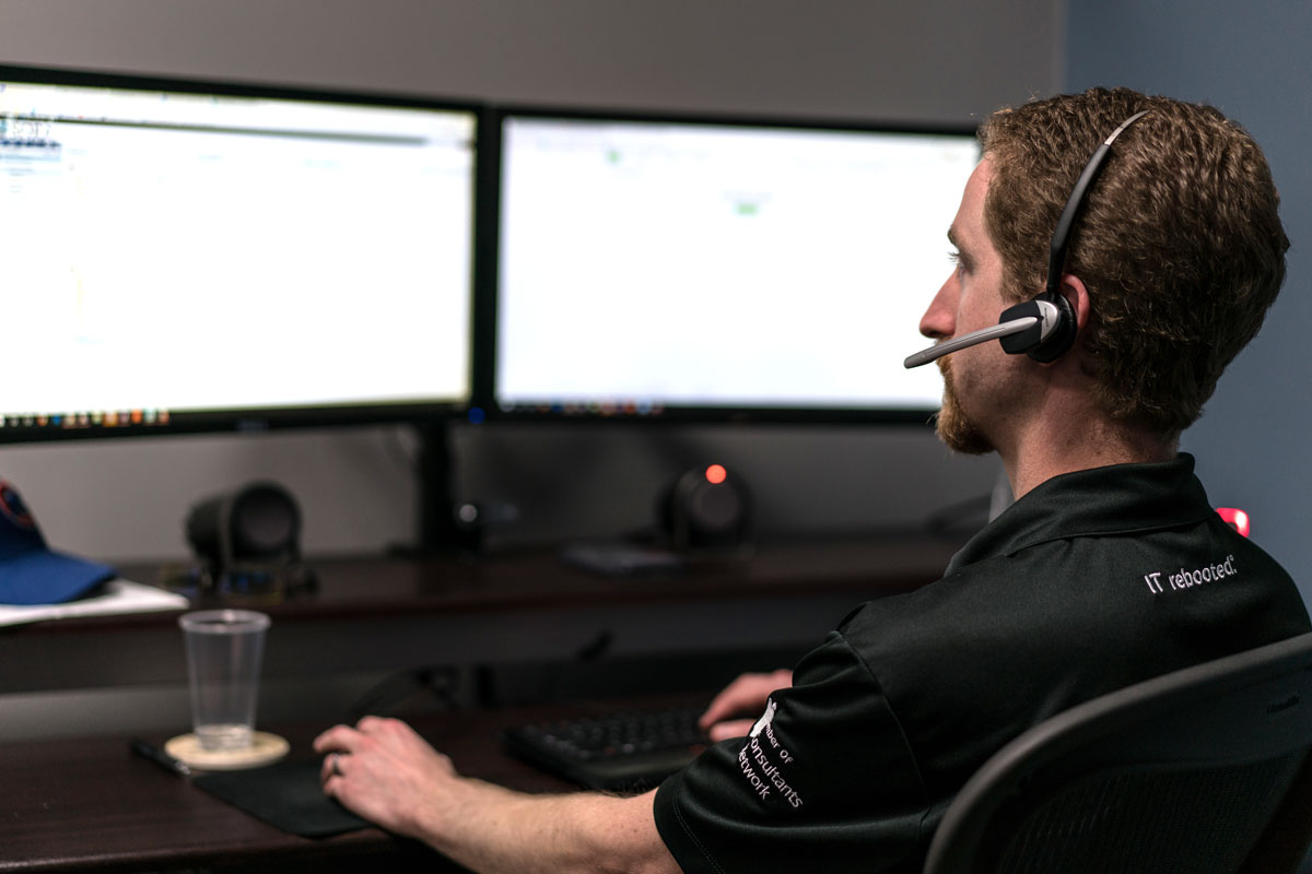 Photo of a IT support service team member with a headset on looking at a computer screen in a Sora's Technology shirt helping a client over the phone.